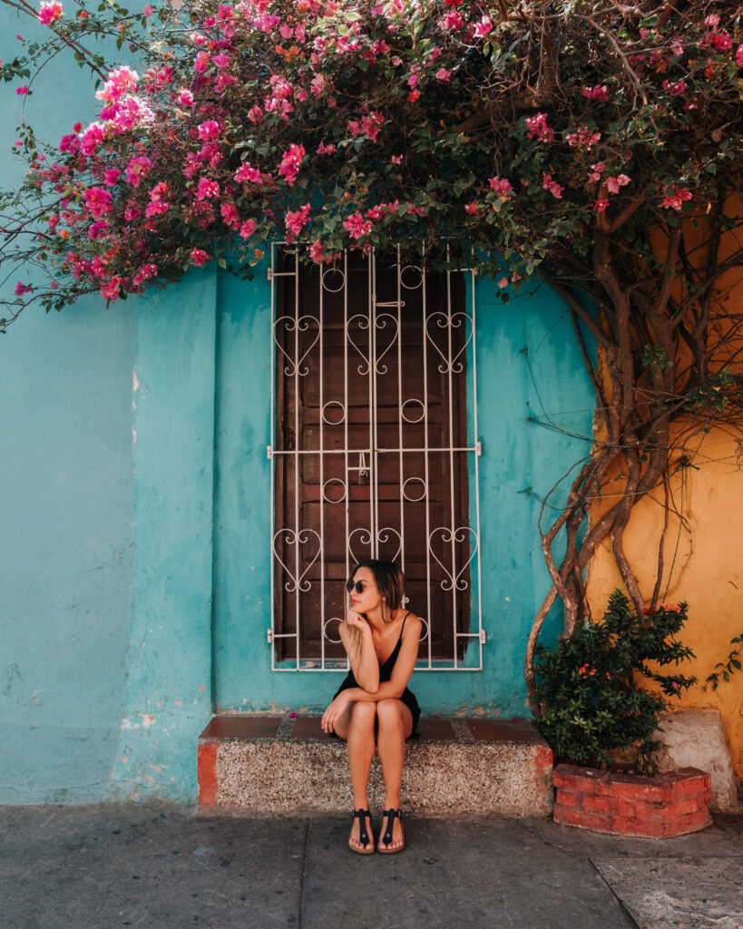 Bree sits on a step under a bougainvillea plant, in front of a bright blue wall on the streets of Medellin