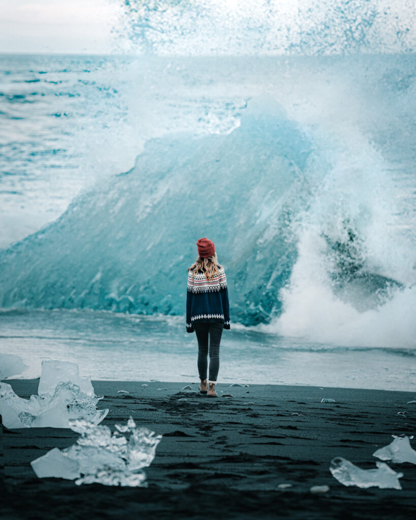 Bree stands on a black sand beach in Iceland looking out at the ocean and a block of ice from a glacier